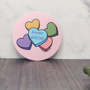 May include: A pink round sticker with four candy hearts in different colors. The hearts are stacked on top of each other. The text "Romance Book Club" is written on the blue heart.