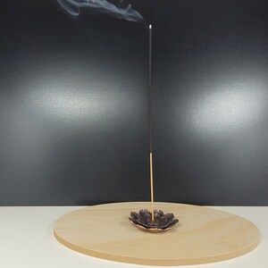 May include: A black incense stick burning in a bronze lotus-shaped incense holder on a wooden base.