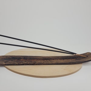 May include: A wooden incense burner with two black incense sticks resting on it. The burner is on a round wooden base.