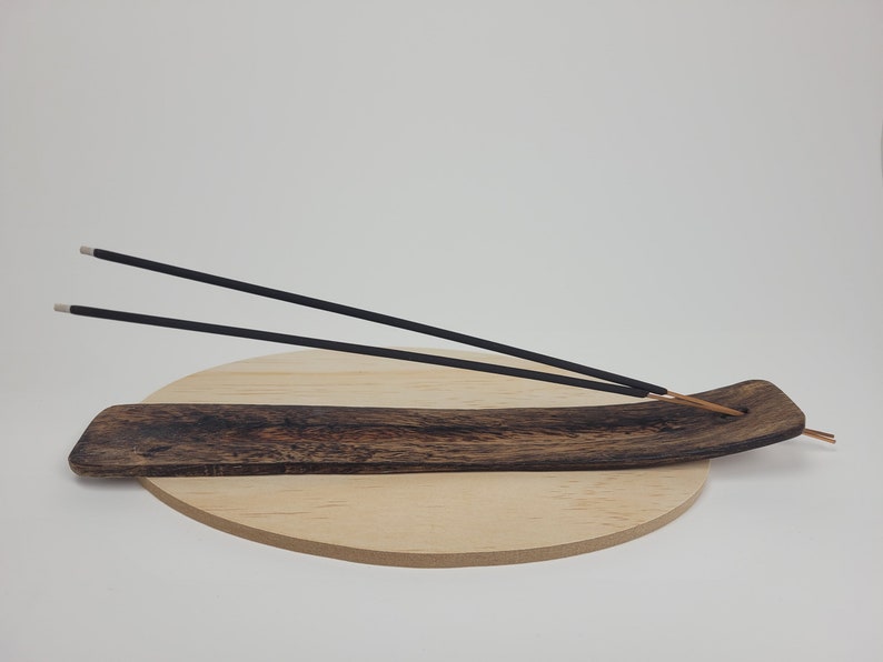 May include: A wooden incense burner with two black incense sticks resting on it. The burner is on a round wooden base.