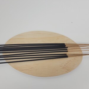 May include: A bundle of black incense sticks with brown wooden tips, arranged on a light brown wooden oval-shaped plate.