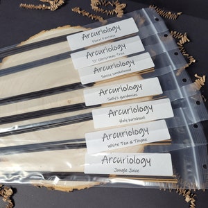 May include: Eight packages of black incense sticks with white labels. The labels have the text "Arcuriology" and the scent name. The scents are Floral Fantasy, "O' Christmas Tree, Sassy Sandalwood, Sally's Gardenias, Holy Patchouli, White Tea & Thyme, and Jungle Juice.