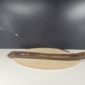 May include: A wooden incense burner with a curved shape and a dark brown finish. A single incense stick is burning and releasing smoke.