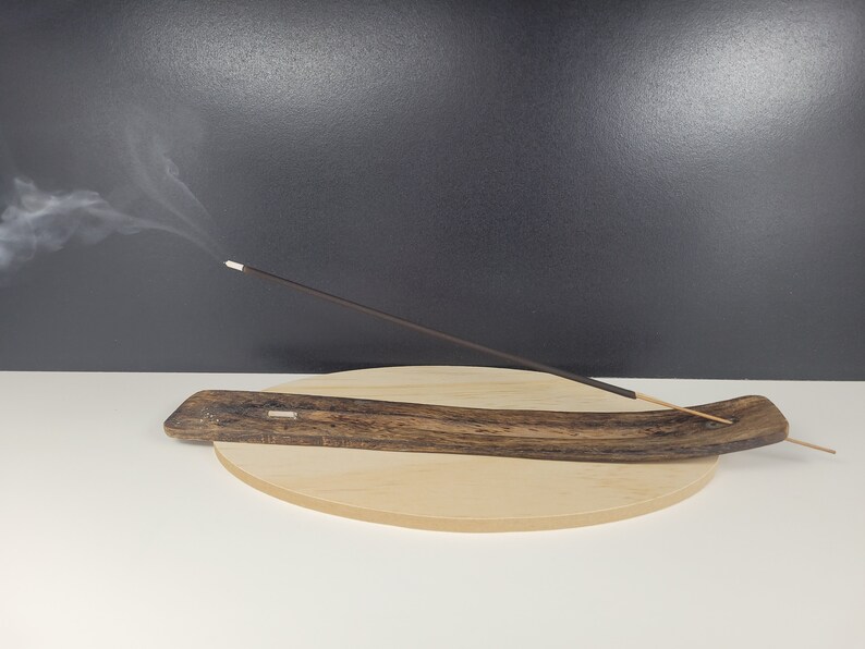 May include: A wooden incense burner with a curved shape and a dark brown finish. A single incense stick is burning and releasing smoke.