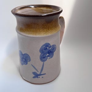 May include: A cylindrical ceramic mug with a light beige body and a dark brown glazed rim. The exterior features hand-painted blue floral motifs consisting of simple brushstrokes. The mug has a sturdy handle on the side and a smooth finish.