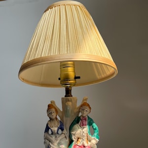 May include: A vintage table lamp with a pleated beige lampshade. The base features a porcelain figurine of three people in period clothing. The figures are painted with blue, green, and pink accents. The lamp has a brass-coloured socket.