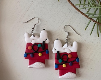May include: A pair of festive earrings featuring a cartoon doghouse design. The doghouse is red with white accents and a string of colorful lights. The dog character is white with black details. The earrings have silver hooks.