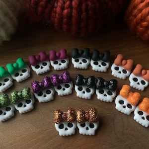 May include: A set of 10 pairs of skull earrings with different colored bows. The bows are made of felt and glitter. The skulls are white and have black eye sockets.