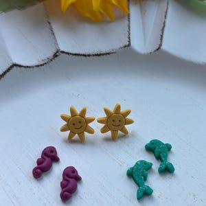 May include: A collection of colorful earrings featuring a summer theme. Includes two pairs of yellow sun-shaped earrings, two purple seahorse earrings, and two green dolphin earrings. The earrings are displayed on a white surface with a yellow sunflower in the background.