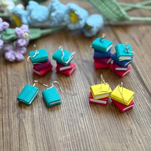 May include: Earrings featuring miniature book designs in teal, yellow, and blue, stacked on red and purple bases. The earrings have silver-colored hooks and are set against a wooden surface with floral accents.