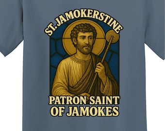 May include: Blue t-shirt featuring a graphic of a saint with a halo, holding a staff. The text reads "ST. JAMOKERSTINE, Patron Saint of Jamokes." The shirt has a crew neck and short sleeves. The FakeSaintShop logo is at the bottom.