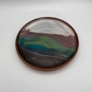 May include: A circular coaster with a smooth, glossy finish featuring a layered color gradient. The design transitions from a dark charcoal base through bands of deep blue, vibrant green, and muted purple, ending in a light gray top section.