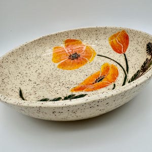 May include: An oval ceramic bowl with a cream-colored base featuring dark brown speckles throughout. The interior is decorated with hand-painted orange poppy flowers and green stems. The bowl has a smooth, glossy finish and a shallow, wide shape.