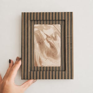 May include: A rectangular picture frame with a striped pattern in shades of brown and black. The frame holds a rectangular image with a marbled design in shades of brown and beige. A hand with painted nails holds the frame.