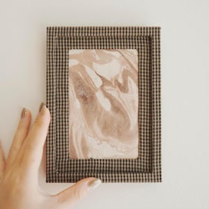 Könnte beinhalten: Ein rechteckiges gerahmtes Kunstwerk mit einem braunen und schwarzen Karomuster. Das Kunstwerk im Inneren zeigt ein beige-braunes Marmormuster. Der Rahmen wird von einer Hand mit lackierten Nägeln gehalten.