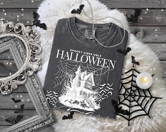 Op de afbeelding: Donkergrijs Halloween-thema T-shirt met de tekst "Spooky Vibes Only Halloween" en een spookhuisafbeelding. Het shirt is omgeven door decoratieve vleermuizen, een spinnenweb en een kaars.