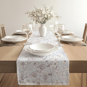 以下が含まれることがあります： 明るい青色の花柄テーブルランナーが敷かれたダイニングテーブル。白いお皿、ワイングラス、カトラリーが各席に配置されています。白い花瓶には白い花のブーケが飾られ、優雅さを添えています。