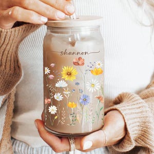 May include: A clear glass reusable water bottle with a bamboo lid and a straw. The bottle has a floral design with yellow, orange, pink, and white flowers. The name "shannon" is written on the bottle.