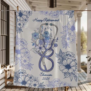Puede incluir: Toalla blanca con diseño floral azul y una ilustración de estetoscopio. El texto "Happy Retirement" está encima del estetoscopio, con "Shannon 2026" debajo. La toalla está colgada en un tendedero.
