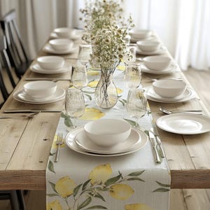 Può includere: Una tavola apparecchiata con piatti, ciotole e bicchieri bianchi. Una tovaglia a tema limoni con limoni gialli e foglie verdi corre al centro. Un vaso trasparente con fiori bianchi è al centro.