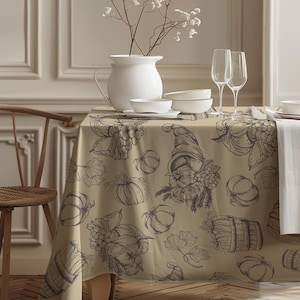 Puede incluir: Un mantel beige con un estampado de temática otoñal con calabazas, uvas y una cornucopia. La mesa está puesta con vajilla blanca, copas de vino y un jarrón blanco con flores.