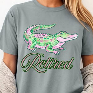 May include: Gray t-shirt featuring a colorful alligator design with pink and green floral patterns. The word "Retired" is written in a cursive font below the alligator.
