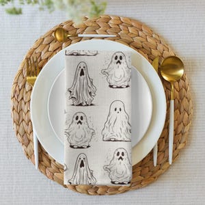 Può includere: Un tovagliolo di stoffa rettangolare con un motivo ripetuto di fantasmi disegnati a mano in bianco e nero su uno sfondo beige chiaro. I fantasmi hanno espressioni e forme varie, disposti in una griglia sulla superficie del tessuto.