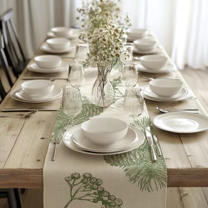 Peut inclure: Une table de salle à manger dressée avec de la vaisselle blanche, des verres transparents et des couverts. Un centre de table floral dans un vase transparent est au centre. Un chemin de table vert et blanc avec des motifs botaniques.