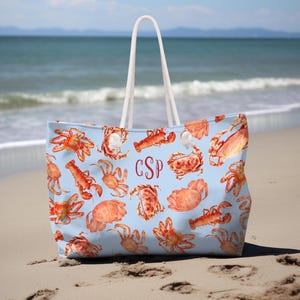 Può includere: Borsa tote azzurra con un motivo ripetuto di granchi e aragoste arancioni. La borsa ha manici in corda bianca e un monogramma bordeaux. La borsa è su una spiaggia sabbiosa con l'oceano sullo sfondo.