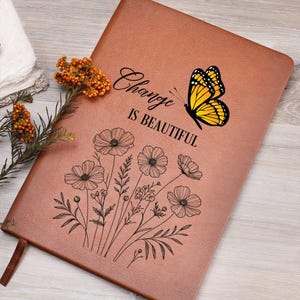 Puede incluir: Un diario de cuero marr&oacute;n con la frase "Change is Beautiful" y una mariposa amarilla. El diario tambi&eacute;n presenta un dise&ntilde;o floral y una cinta de marcador marr&oacute;n. Una ramita de flores naranjas descansa sobre el diario.
