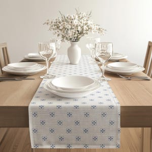 Könnte beinhalten: Ein Esstisch, der für eine Mahlzeit gedeckt ist, mit einem weißen Blumengesteck in einer weißen Vase, weißen Tellern und Weingläsern. Ein gemusterter Tischläufer mit blauen Blumendesigns schmückt den Holztisch.