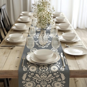 May include: A dining table set for a meal. White plates, bowls, and glasses are arranged on a gray runner with a floral design. A clear vase with flowers sits in the center. The table is made of light-colored wood.