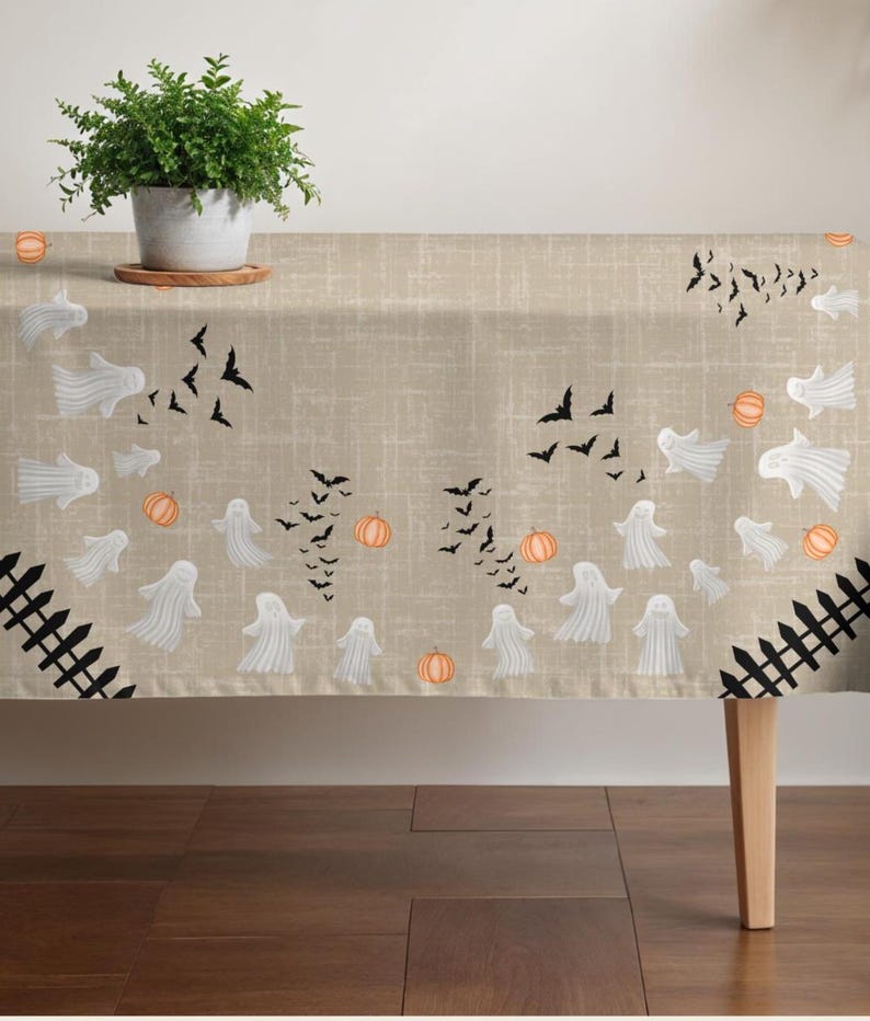 K&ouml;nnte beinhalten: Eine beigefarbene Tischdecke mit strukturierter Stoffoptik, die ein wiederkehrendes Halloween-Muster aus wei&szlig;en Geistern, schwarzen fliegenden Flederm&auml;usen, orangefarbenen K&uuml;rbissen und schwarzen Zaunabschnitten an den Ecken zeigt.