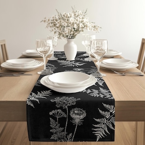 May include: A black table runner with a white botanical design of ferns and wildflowers. The runner is on a wooden table, set with white plates, glasses, and cutlery. A white vase holds white flowers.