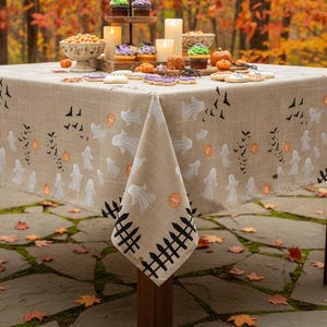 K&ouml;nnte beinhalten: Eine quadratische, beige Tischdecke mit einem wiederkehrenden Halloween-Muster aus wei&szlig;en Geistern, orangefarbenen K&uuml;rbissen, schwarzen Flederm&auml;usen und einer schwarzen Zaunbord&uuml;re am unteren Rand. Der Stoff wirkt strukturiert und bedeckt einen Holztisch.
