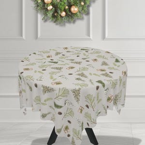 Pode incluir: Uma toalha de mesa redonda cinza claro com um padrão festivo de Natal. O design inclui galhos de pinheiro verdes, pinhas e presentes embrulhados. A toalha de mesa está sobre uma mesa preta.