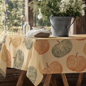 May include: A beige tablecloth featuring a pumpkin pattern in orange, green, and yellow. The table is set with white plates, a folded napkin, and a metal bucket filled with white flowers. The table is made of wood.