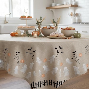 K&ouml;nnte beinhalten: Eine runde beige Tischdecke mit einem sich wiederholenden Muster aus wei&szlig;en Geistern, orangefarbenen K&uuml;rbissen und schwarzen Flederm&auml;usen, mit einem schwarzen Lattenzaun-Design am unteren Rand. Der Stoff hat eine strukturierte Optik und bedeckt einen runden Holztisch.