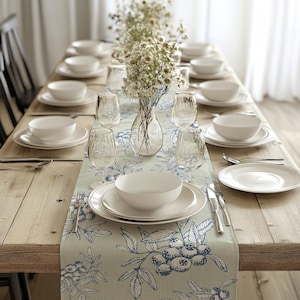 May include: A dining table set for a meal, featuring white dinnerware, clear glassware, and a floral centerpiece. A blue and white patterned table runner adds a decorative touch. The table is made of light-colored wood.