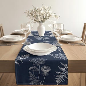 Puede incluir: Camino de mesa azul marino con un estampado botánico blanco de helechos y flores silvestres. El camino de mesa está colocado sobre una mesa de madera, complementado con vajilla blanca, cristalería y un jarrón de flores blancas, creando un ambiente elegante.