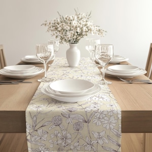 May include: A dining table set with white plates, wine glasses, and cutlery. A floral table runner with a cream background and white and purple botanical illustrations runs down the centre. A white vase holds a bouquet of white flowers.