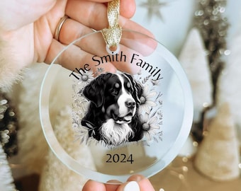 Op de afbeelding: Een rond, transparant acryl ornament met een zwart-witte illustratie van een Berner Sennenhond, omringd door florale lijntekeningen. De tekst luidt The Smith Family en 2024. Het bevat een gouden lintlus om aan de boom te hangen.