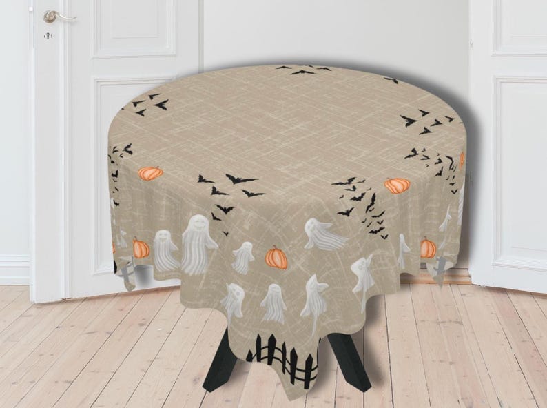 K&ouml;nnte beinhalten: Eine runde, beige Tischdecke mit strukturierter Leinenoptik, die mit einem wiederkehrenden Halloween-Muster aus wei&szlig;en Geistern, orangefarbenen K&uuml;rbissen, schwarzen Flederm&auml;usen und einer schwarzen Zaunbord&uuml;re am unteren Rand bedruckt ist.