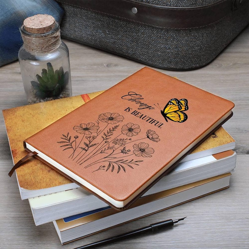 Puede incluir: Una pila de cuadernos. El cuaderno superior tiene una cubierta de cuero marr&oacute;n con un dise&ntilde;o floral, una mariposa amarilla y el texto "Change is Beautiful". Se ve un frasco de vidrio con tapa de corcho y una planta peque&ntilde;a.