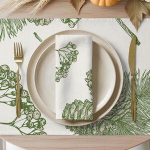 Puede incluir: Una mesa puesta con un plato blanco, una servilleta y un mantel individual. La servilleta y el mantel individual presentan un diseño botánico verde de piñas y bayas. Un tenedor y un cuchillo dorados están a ambos lados del plato.