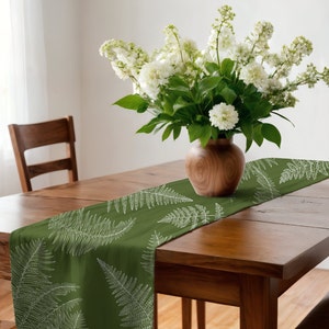 May include: A green table runner with a white fern pattern. The runner is on a wooden table with a vase of white flowers in the center.