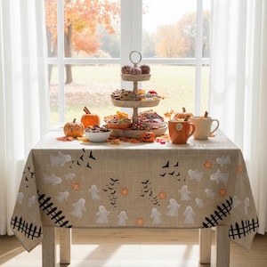K&ouml;nnte beinhalten: Eine rechteckige beige Tischdecke mit einem wiederkehrenden Muster aus wei&szlig;en Geistern, schwarzen Flederm&auml;usen, orangefarbenen K&uuml;rbissen und schwarzen Lattenzaunabschnitten. Der Stoff hat eine sichtbare Webstruktur und liegt auf einem Tisch.