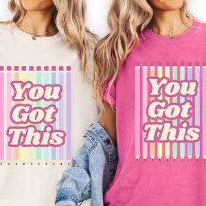 May include: Two t-shirts, one cream and one pink, each with the text "You Got This" in a retro font. The design includes vertical pastel-colored pencil illustrations. The shirts are short-sleeved.