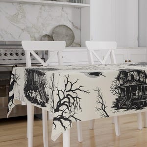 May include: A rectangular tablecloth with a spooky Halloween theme. The off-white cloth features a black and white print of haunted houses, bare trees, and a full moon. The tablecloth is draped over a white table.