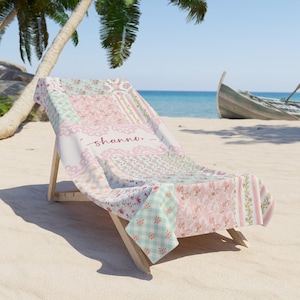 Puede incluir: Una toalla de playa floral rosa y verde con el nombre "shanna" escrito en ella. La toalla está doblada y colocada en una silla de playa de madera.